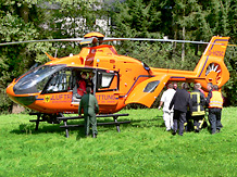 Sanitter und Feuerwehrleute tragen den Verletzten zu dem auf einer Wiese stehenden orangen Rettungshubschrauber
